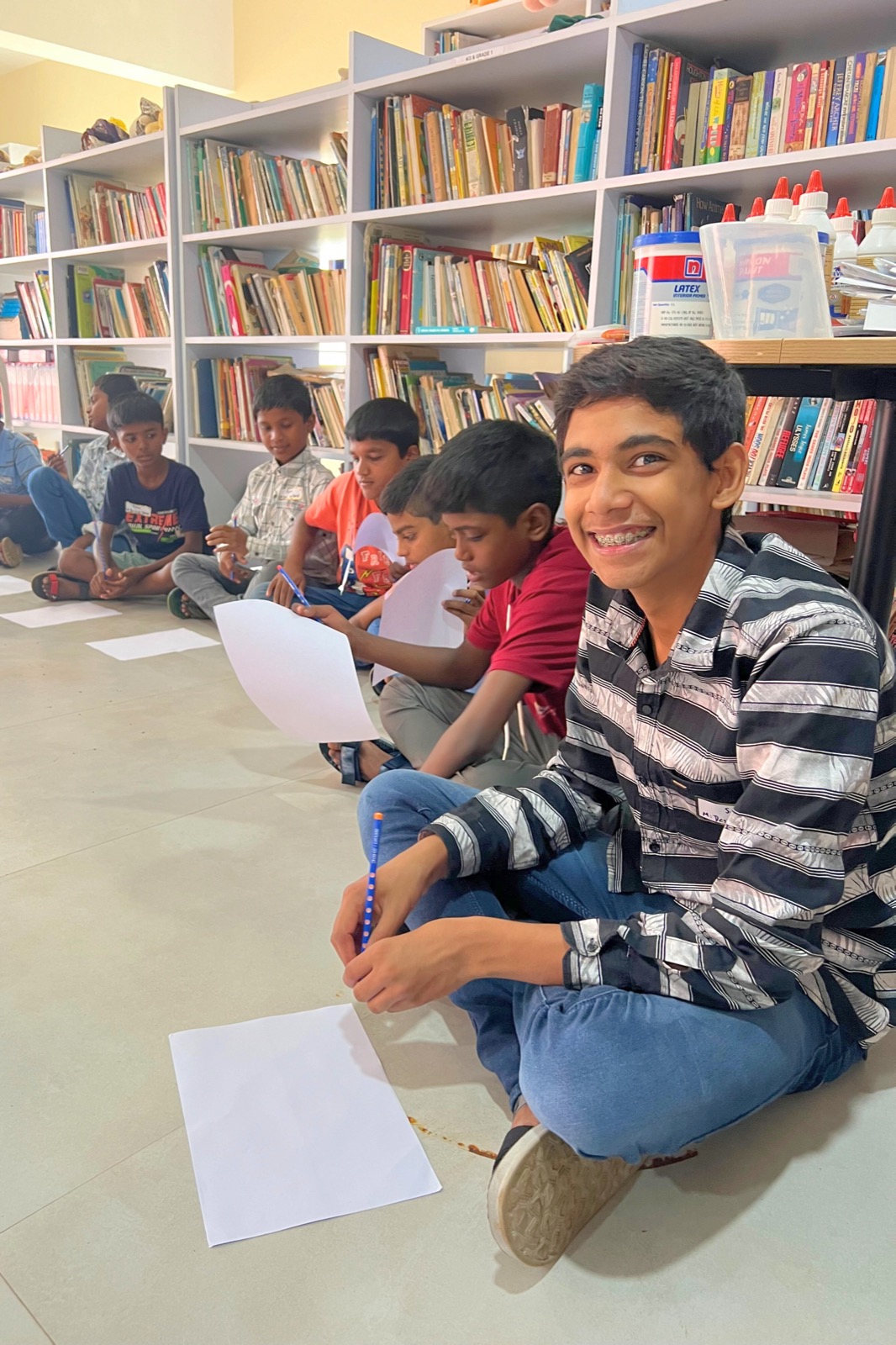 Students sitting on a library floor writing, one smiling at the camera