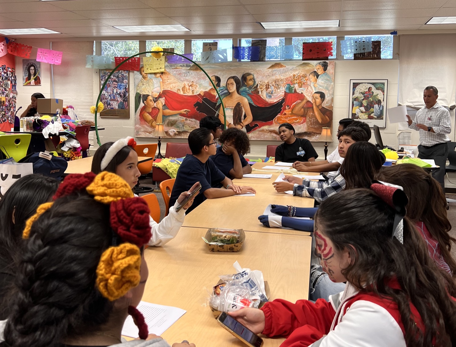Students seated around tables in an art classroom with a large mural behind them